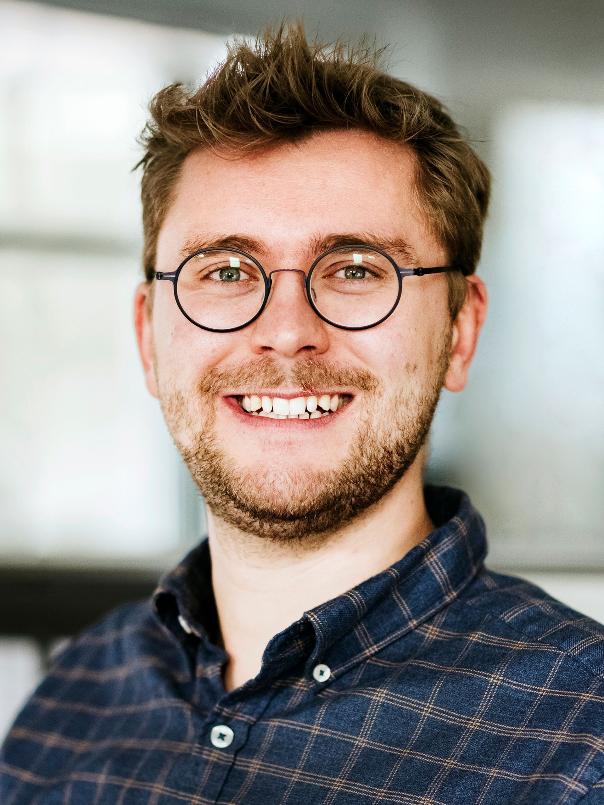 Eine lächelnde Person mit kurzen braunen Haaren und runder Brille, die ein dunkelblau kariertes Hemd mit Knöpfen trägt.