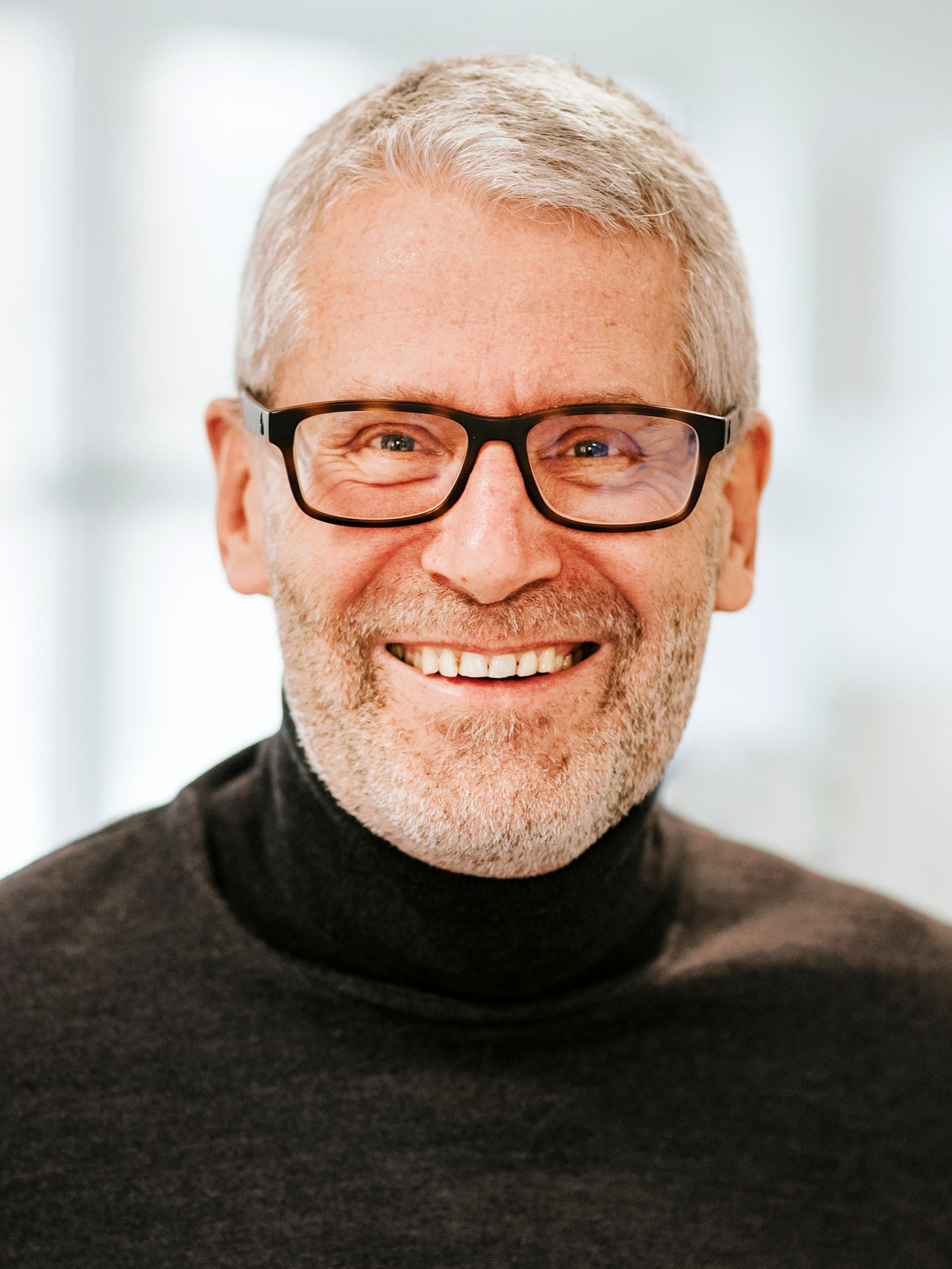 Eine lächelnde Person mit kurzen grauen Haaren und Brille, die einen dunklen Rollkragenpullover trägt,