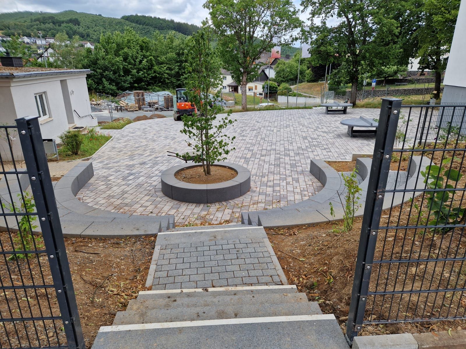 Über eine Betontreppe gelangt man hinunter zu einem gepflasterten Außenbereich mit einem zentralen runden Pflanzbeet, 