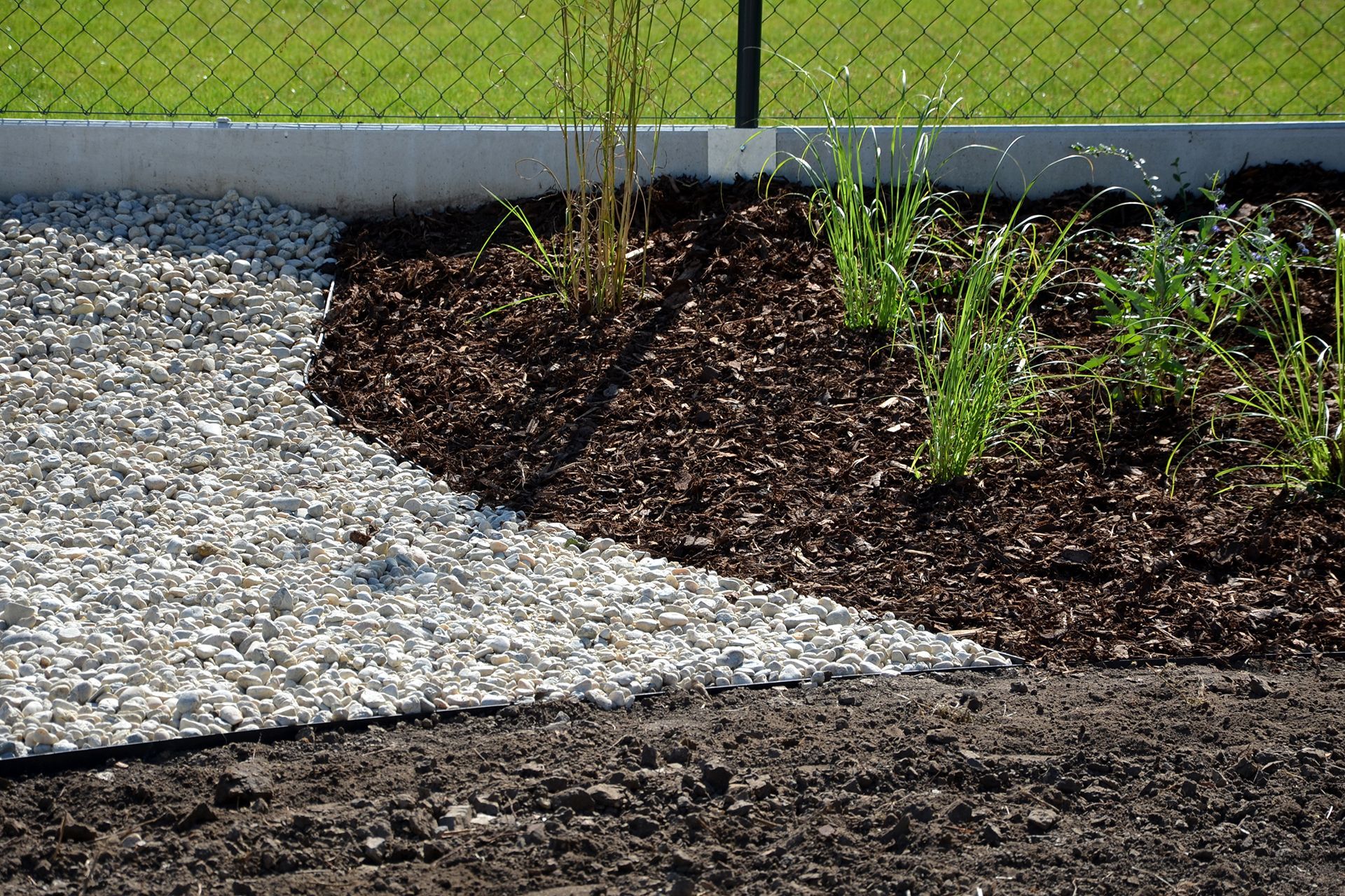 a garden with gravel , mulch and plants in front of a chain link fence .