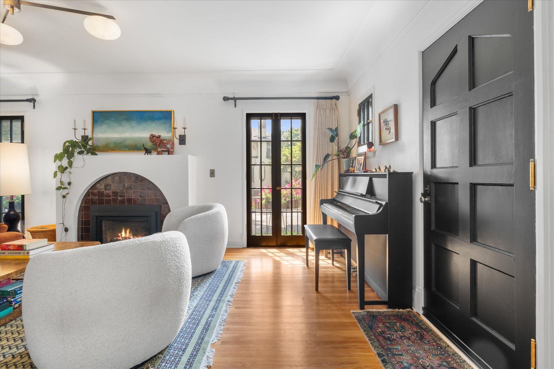 A living room with white walls, a brick fireplace, two fuzzy white chairs, a black piano, and a large black front door.