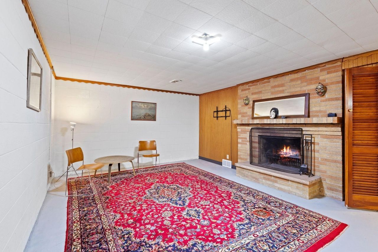 A basement living area with a large red patterned rug, a stone fireplace, and two wooden chairs.