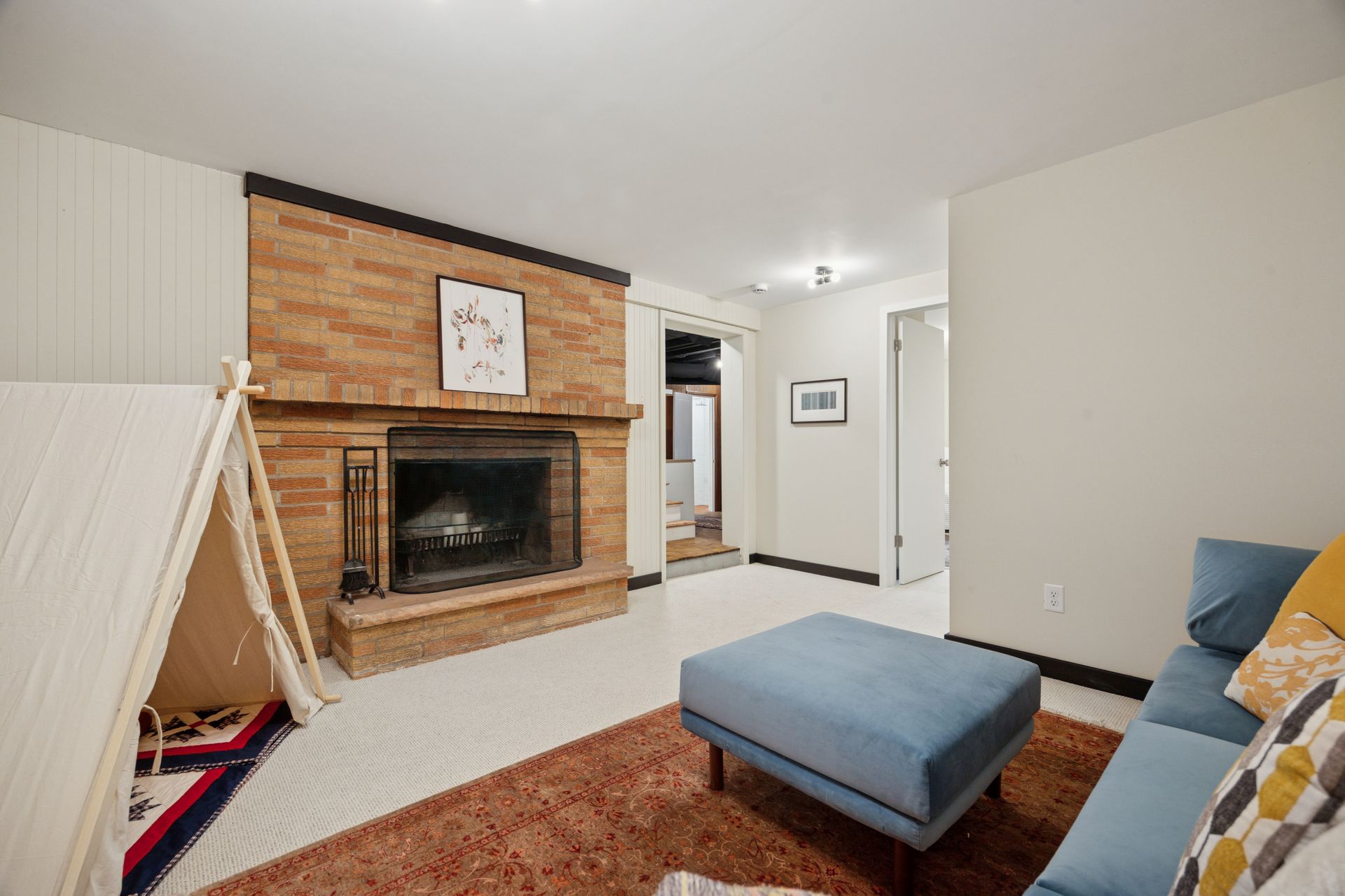 Living room with a brick fireplace, a teepee, a blue ottoman, and a sofa on a brown rug against light walls.