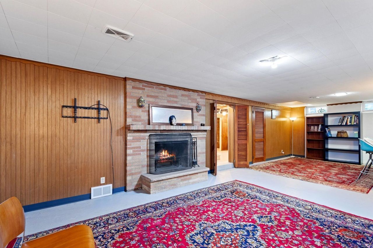 A basement living area with wood-paneled walls, a brick fireplace, two large red Persian rugs, and a tall black bookcase.
