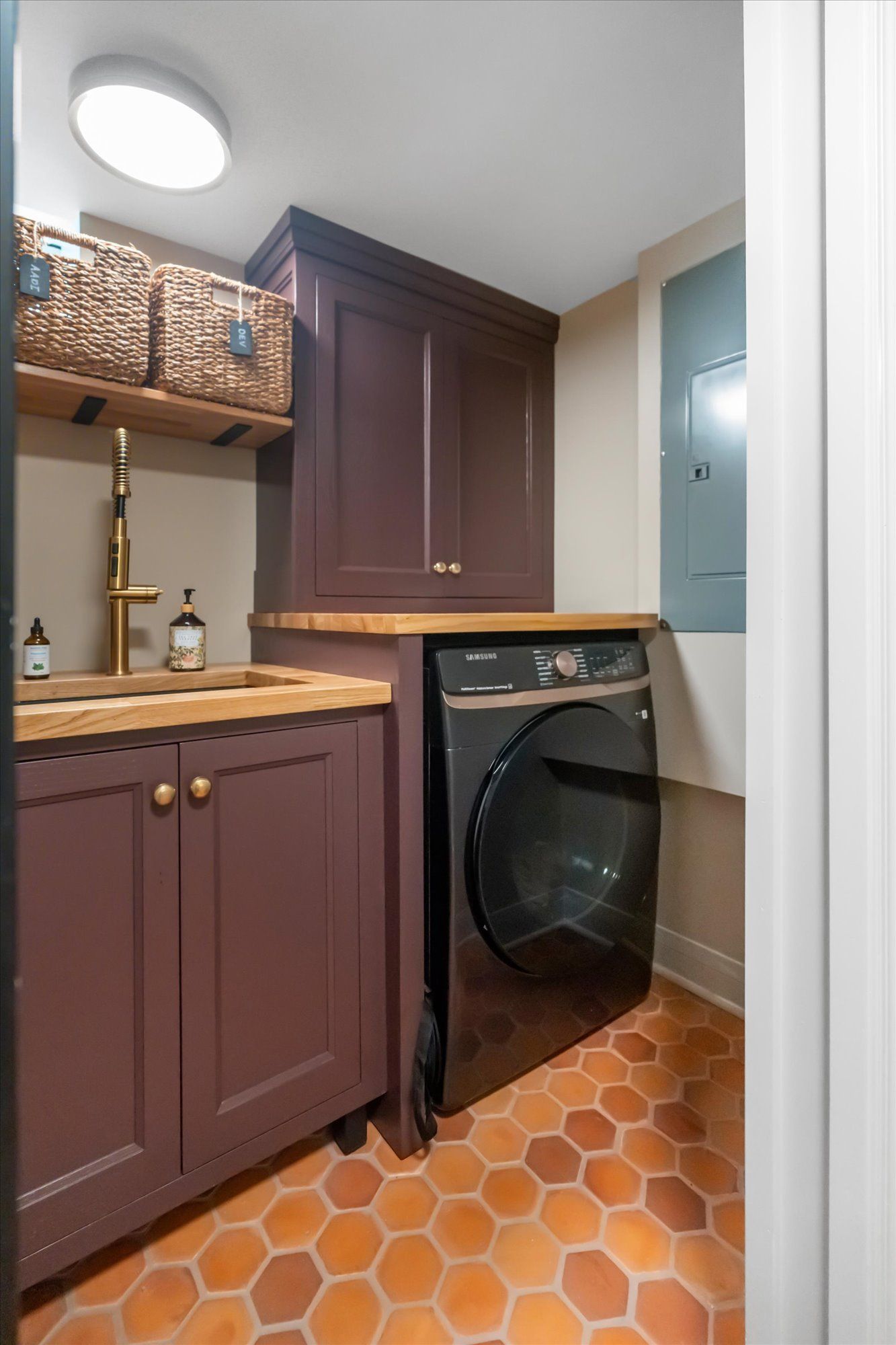 A laundry room with dark plum cabinets, a wood countertop with a sink, a black washer, and orange hexagonal floor tiles.