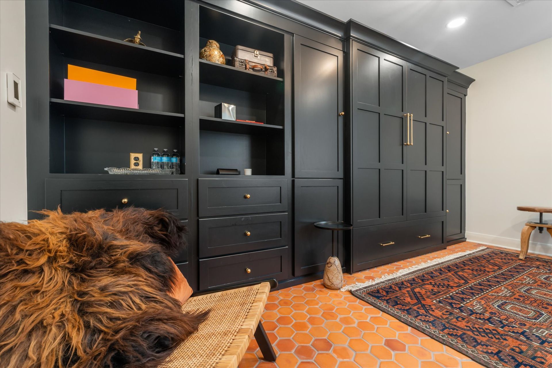 Black cabinetry with open shelving and a hidden wall bed, set in a room with a hexagonal tile floor and a patterned rug.
