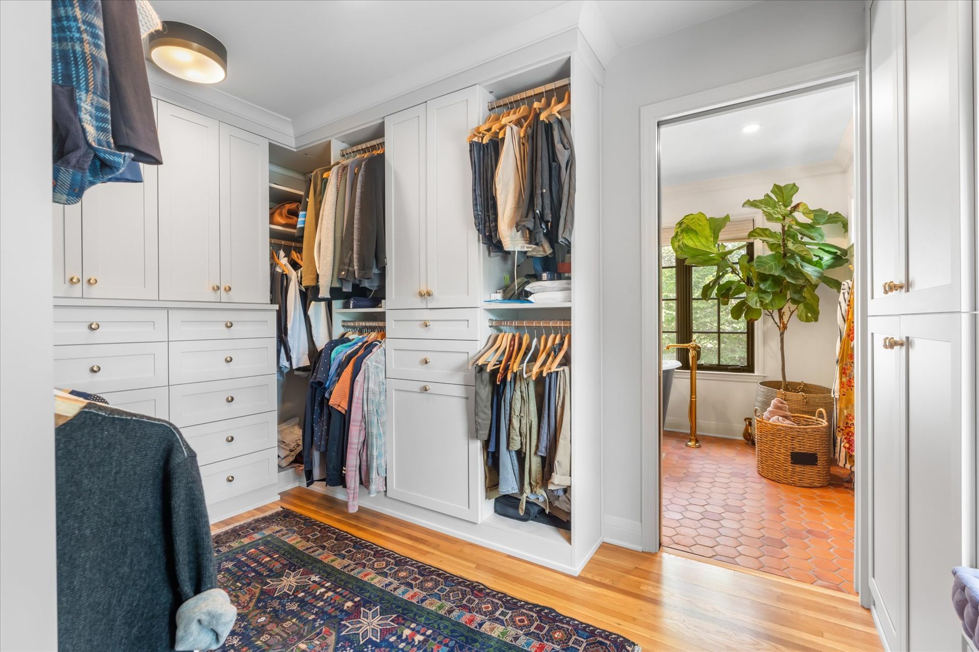 A well-lit walk-in closet featuring white cabinetry, hanging clothes, and a patterned rug, leading into a bathroom.