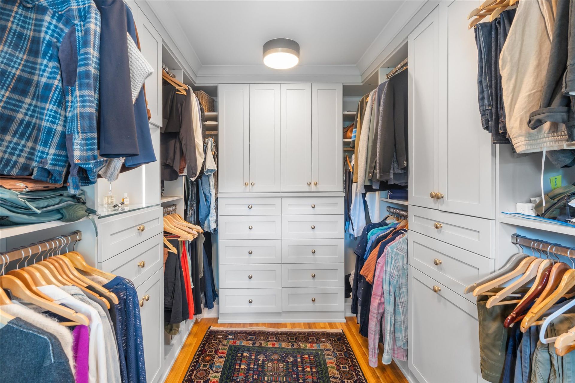 A walk-in closet with white cabinets, drawers, hanging clothes on both sides, and a patterned rug on a wooden floor.