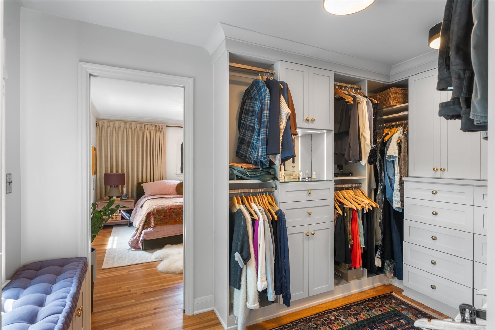 A well-organized walk-in closet with grey cabinetry, hanging clothes, and a bench, leading to a bedroom.
