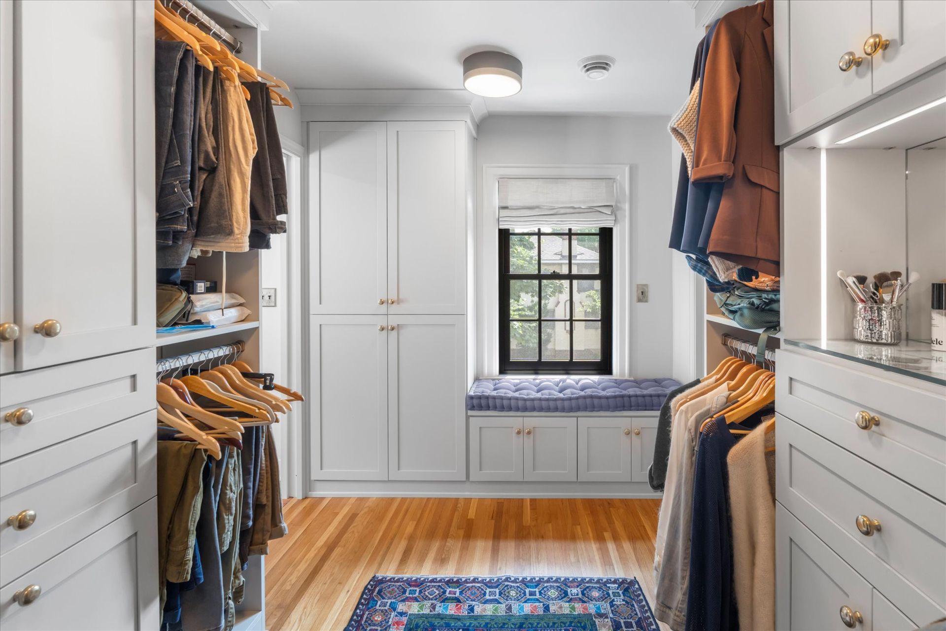 A light-filled walk-in closet featuring white cabinetry, hanging clothes, hardwood floors, and a window seat.