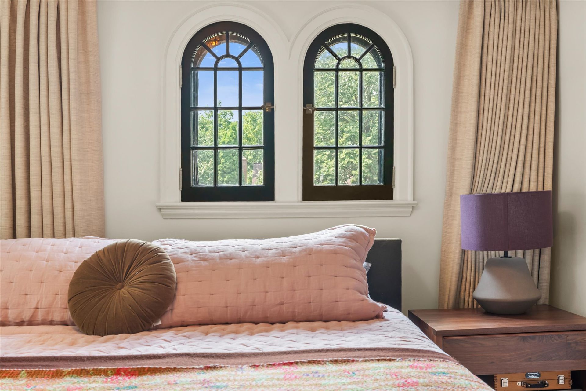 A bedroom with a pink quilted bed, a circular brown pillow, twin arched windows, beige curtains, and a purple lamp.