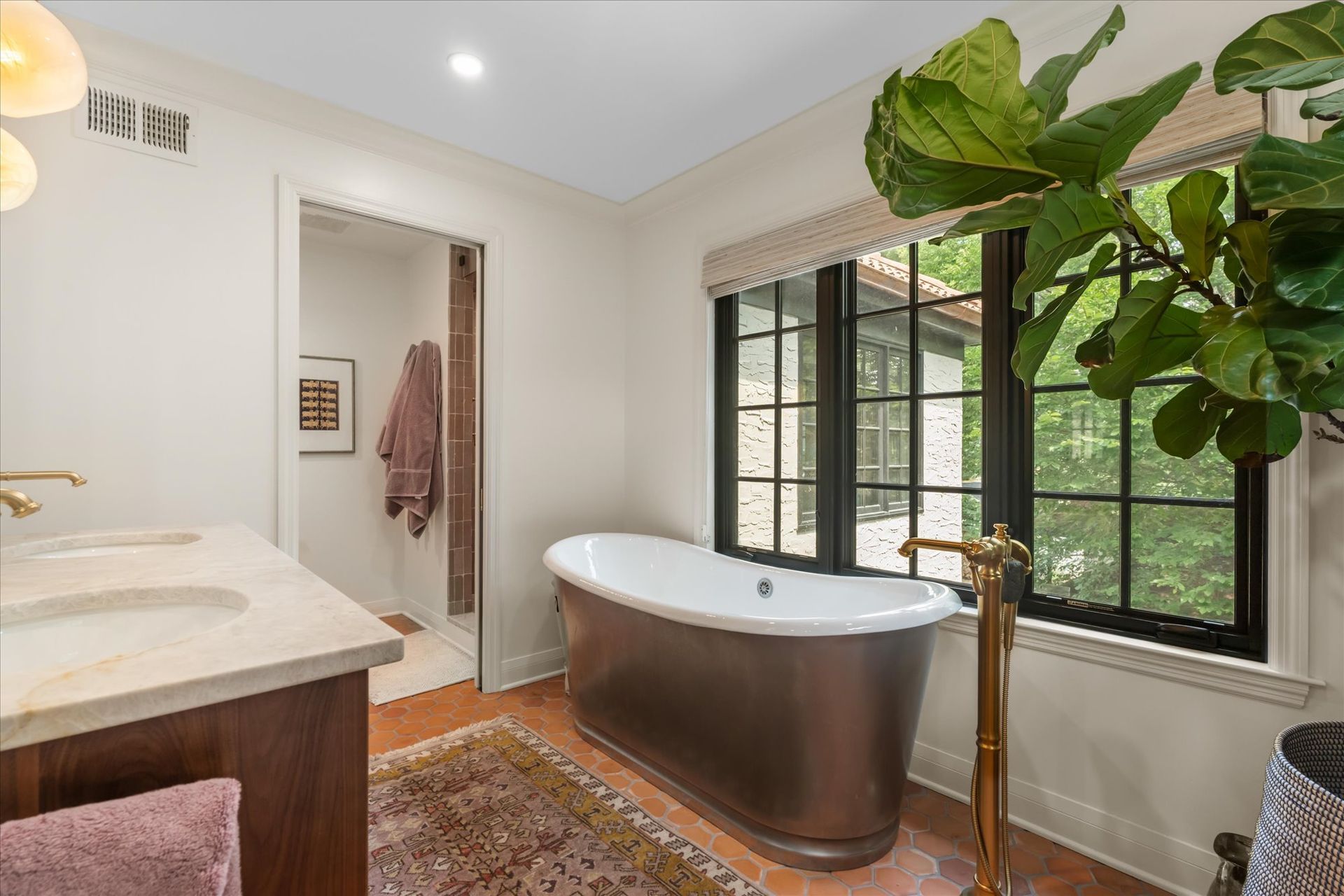 A bathroom features a bronze pedestal bathtub, a dark wood vanity, a window with dark frames, and a potted plant.