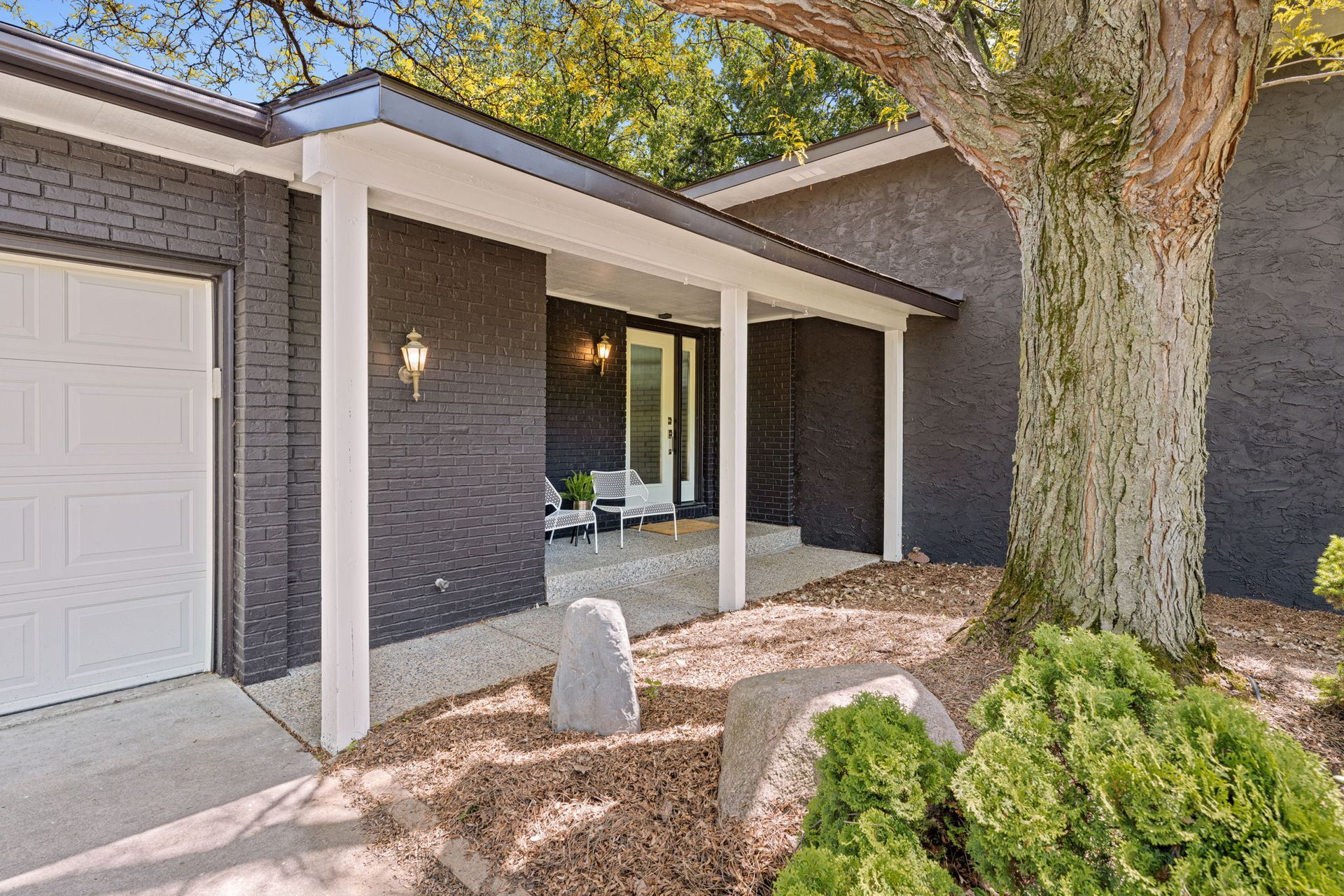 Front entry of a brick house with white trim, garage door, porch light, and a large tree in the yard.