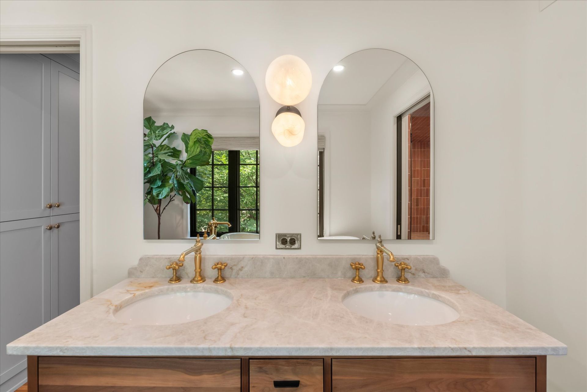 A double vanity with marble countertop, brass faucets, two arched mirrors, and modern spherical wall lighting.
