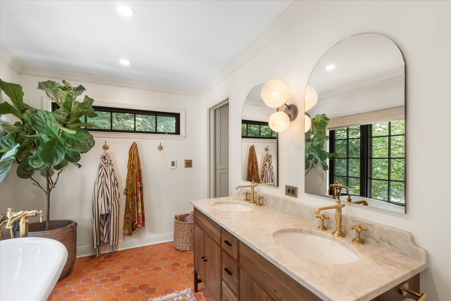 A bright bathroom with terracotta floors, a wood vanity, gold fixtures, two arched mirrors, and a large fiddle leaf fig.