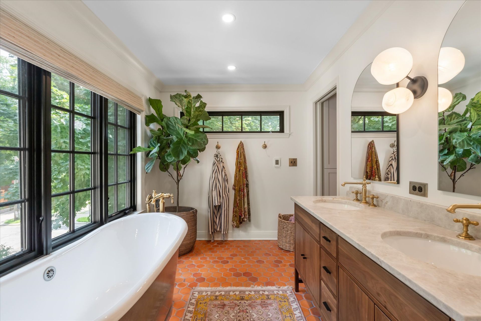 A bright, modern bathroom with a white freestanding tub, double vanity, large windows, a fiddle leaf fig, and terracotta floor.