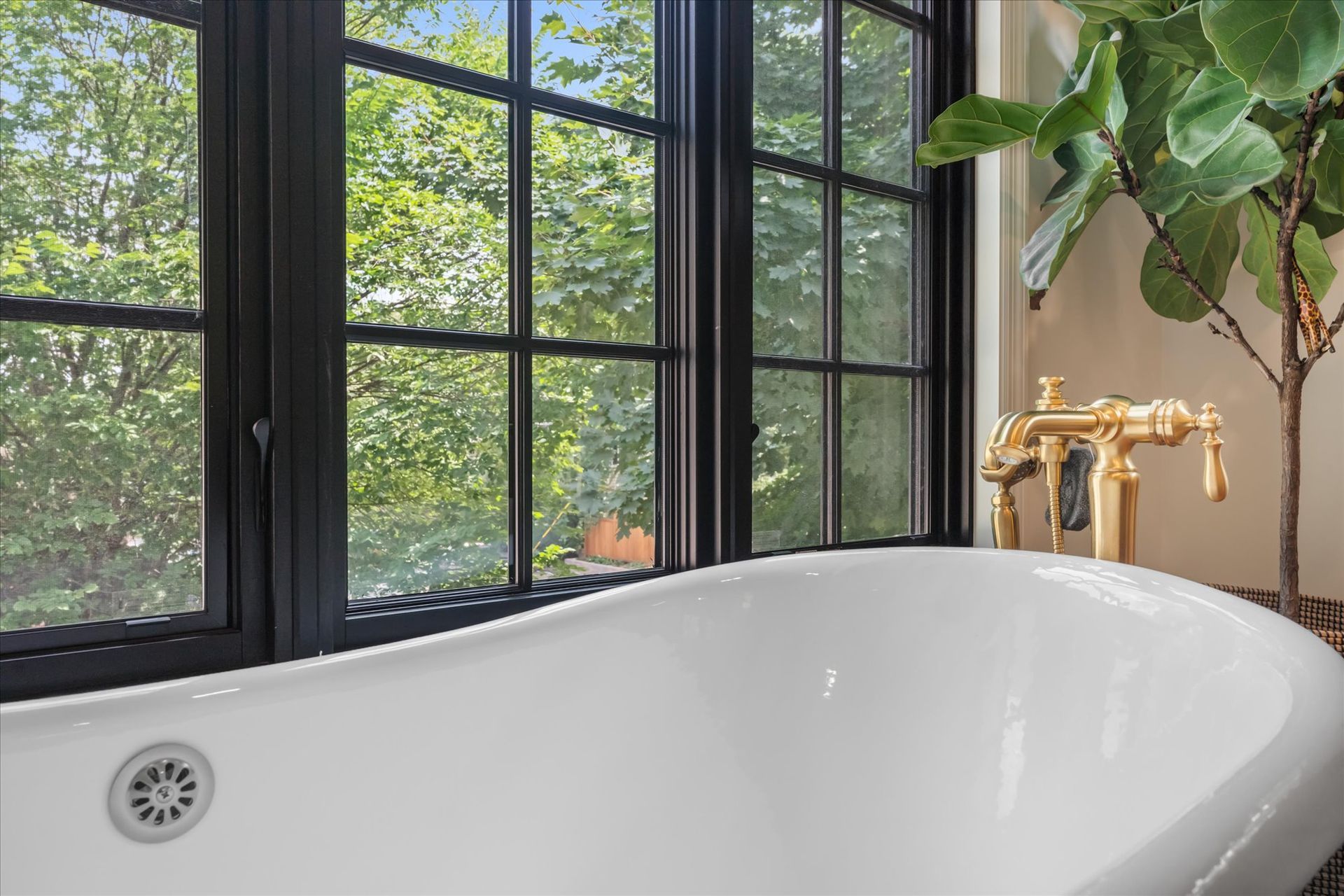 White freestanding bathtub with gold fixtures positioned in front of a window with black frames and a view of trees.