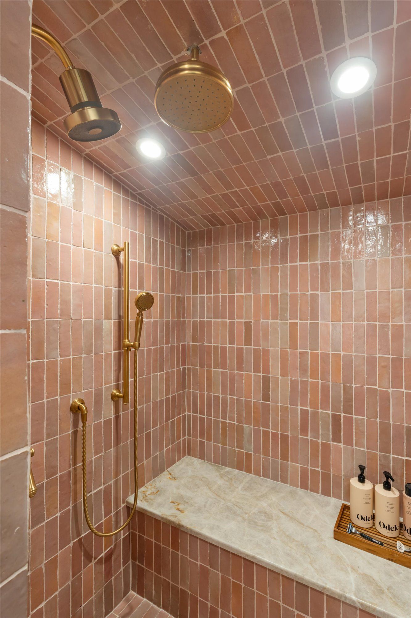 A tiled shower with a marble bench, brass fixtures, a rainfall showerhead, and matching handheld shower.