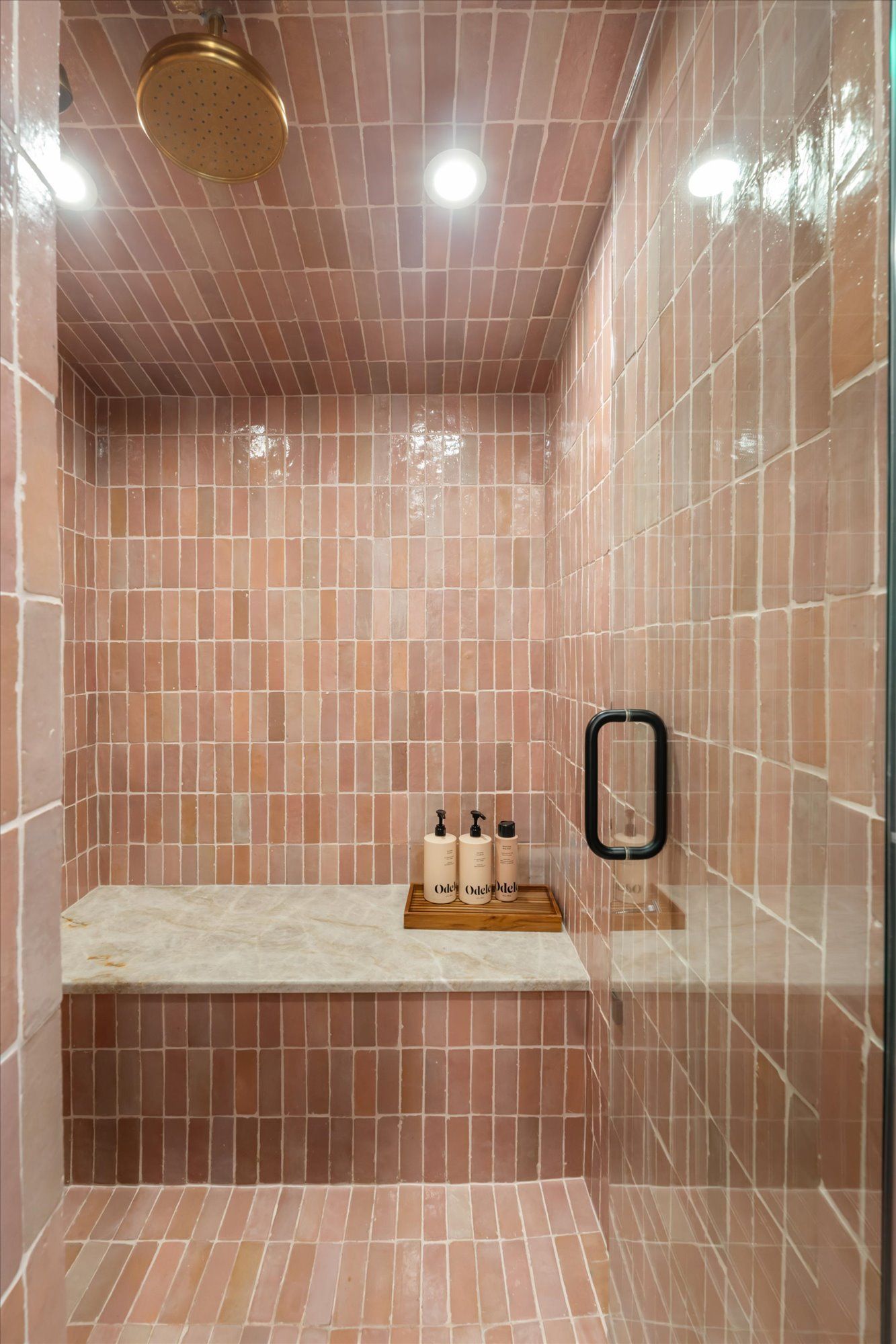 A shower with pinkish-tan rectangular wall and floor tiles, a stone bench, and a gold rainfall showerhead.
