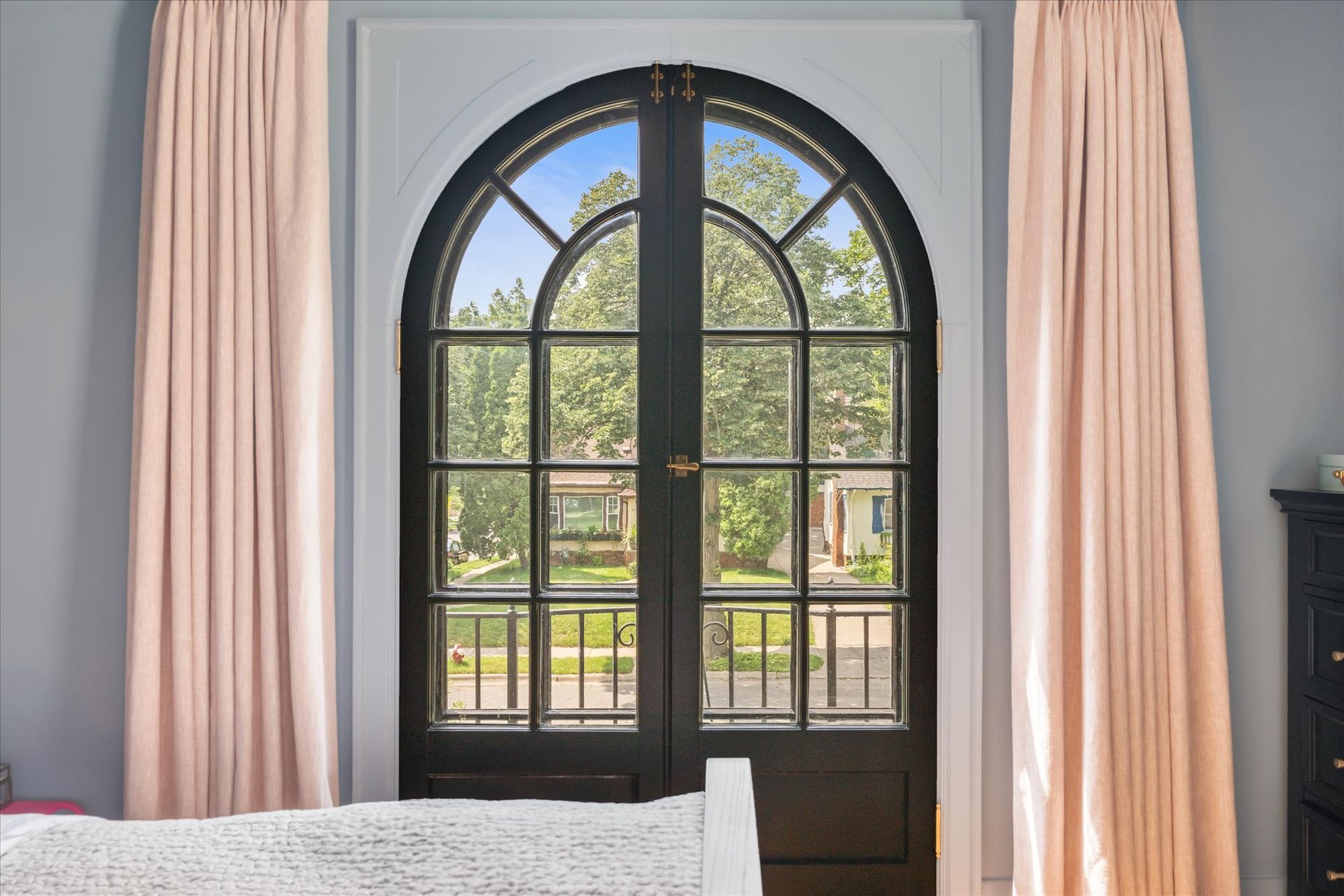 Arched black-framed French doors with clear glass panes open to a view of trees, flanked by light pink curtains.