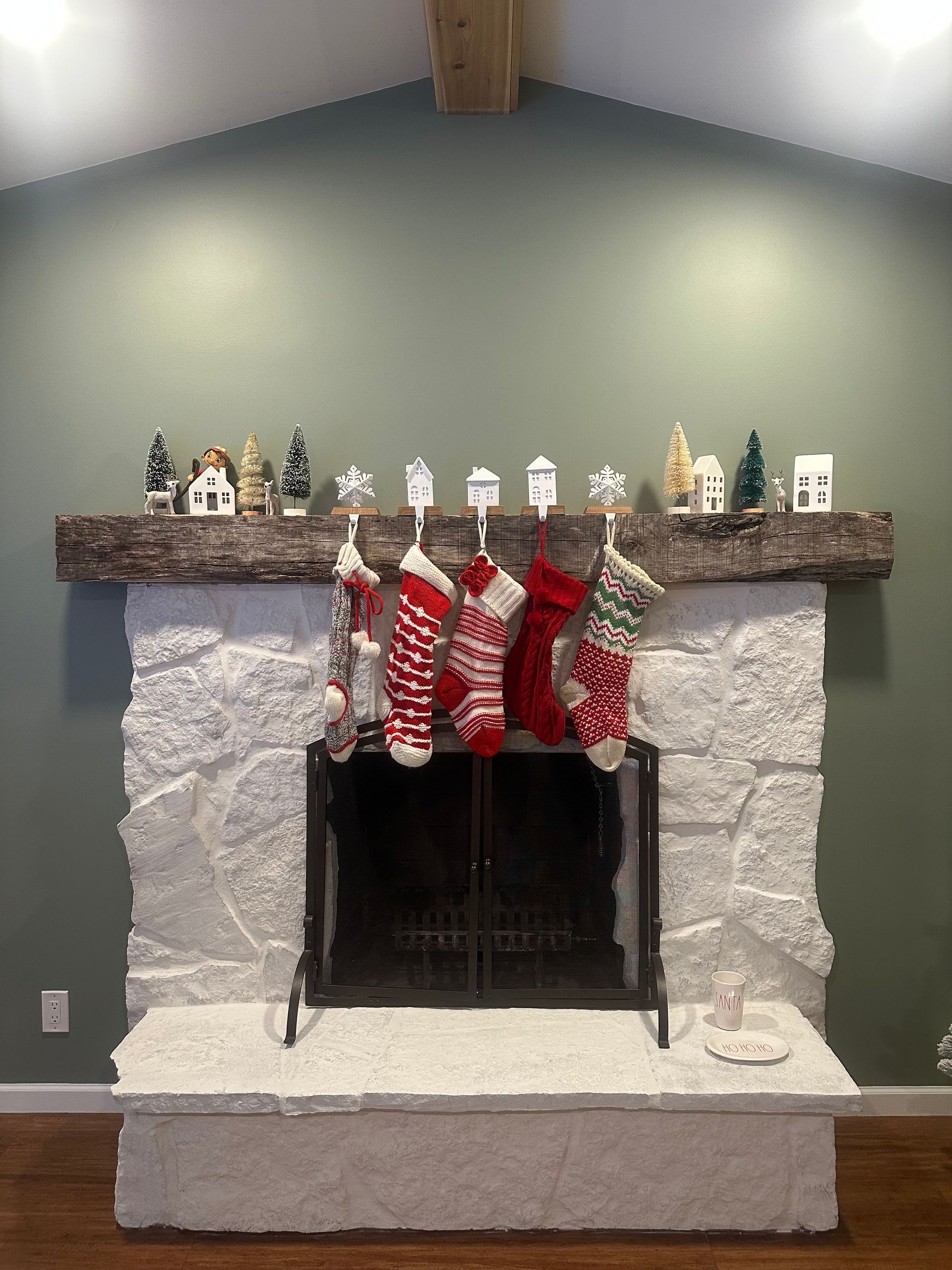 A white stone fireplace with a wooden mantel decorated with miniature houses, trees, and five hanging Christmas stockings.