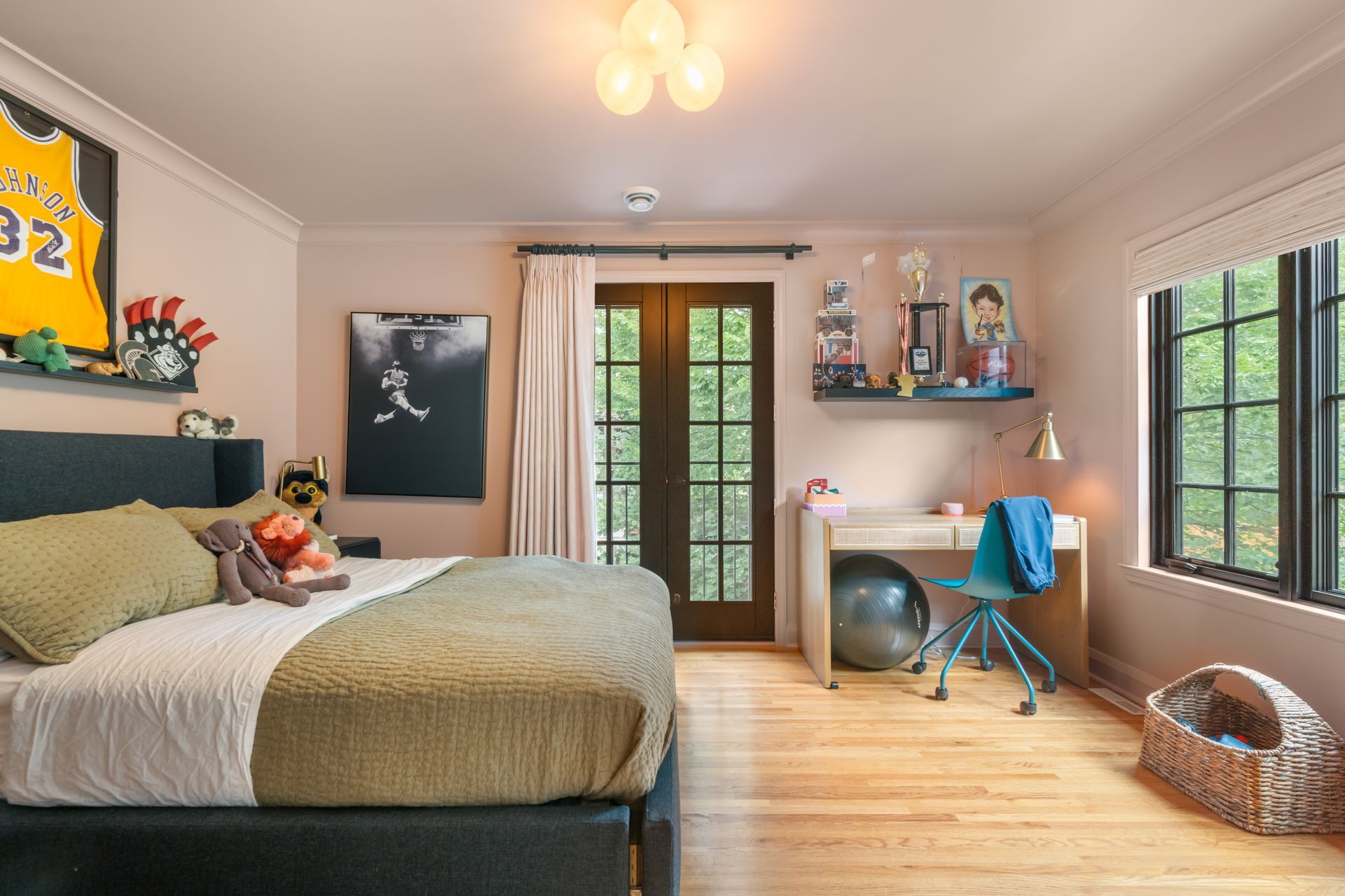 A bedroom with a bed, desk, chair, exercise ball, large window, and balcony door. Neutral walls and hardwood floors.