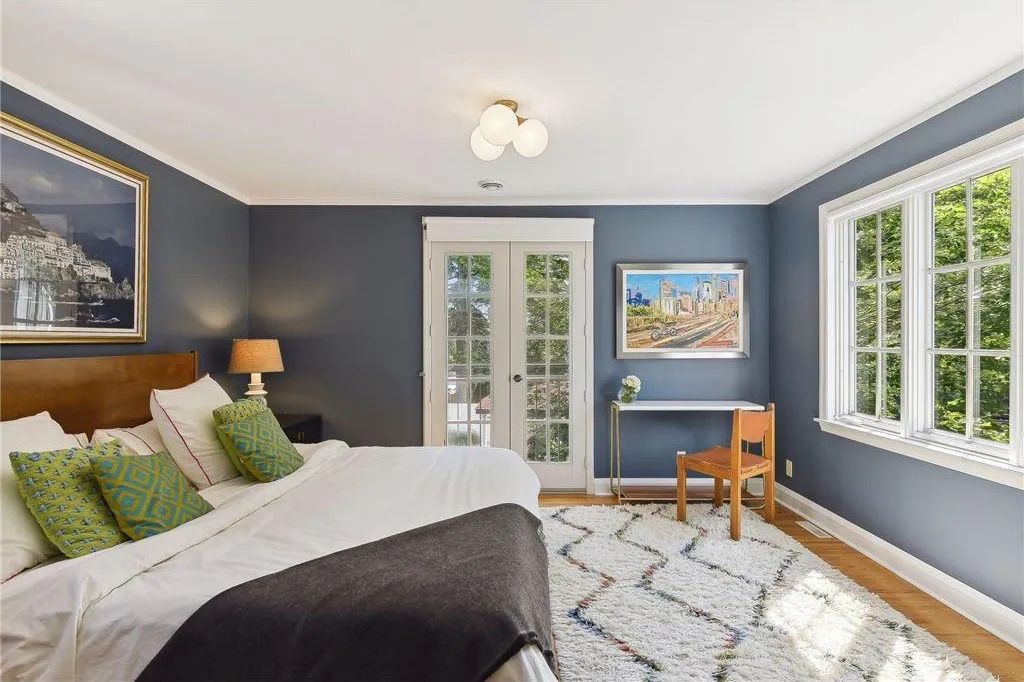 A bedroom with blue walls, a large bed with a dark throw, a white textured rug, glass doors, and a small desk with a chair.