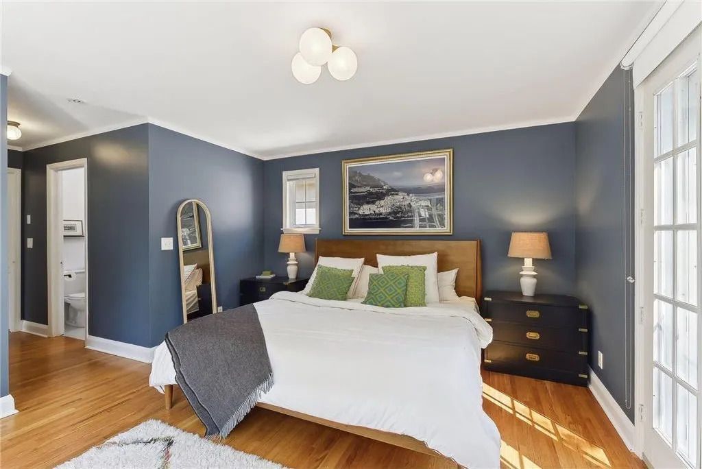 A bedroom with dark blue walls, a white bed, wood floors, a framed picture, and two lamps on bedside tables.