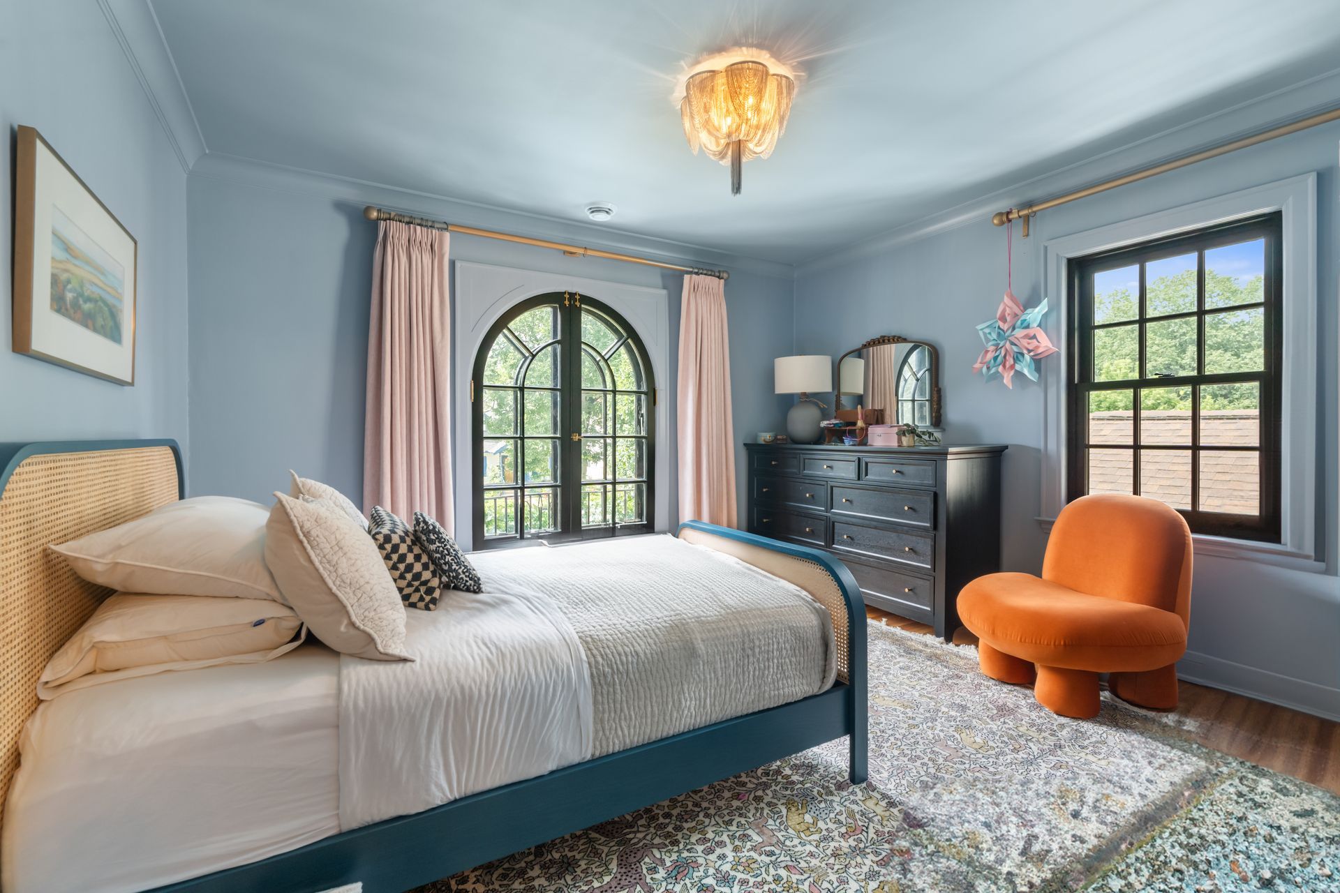 A bedroom with light blue walls, a bed with tan bedding, a dark dresser, an orange armchair, and a window with curtains.