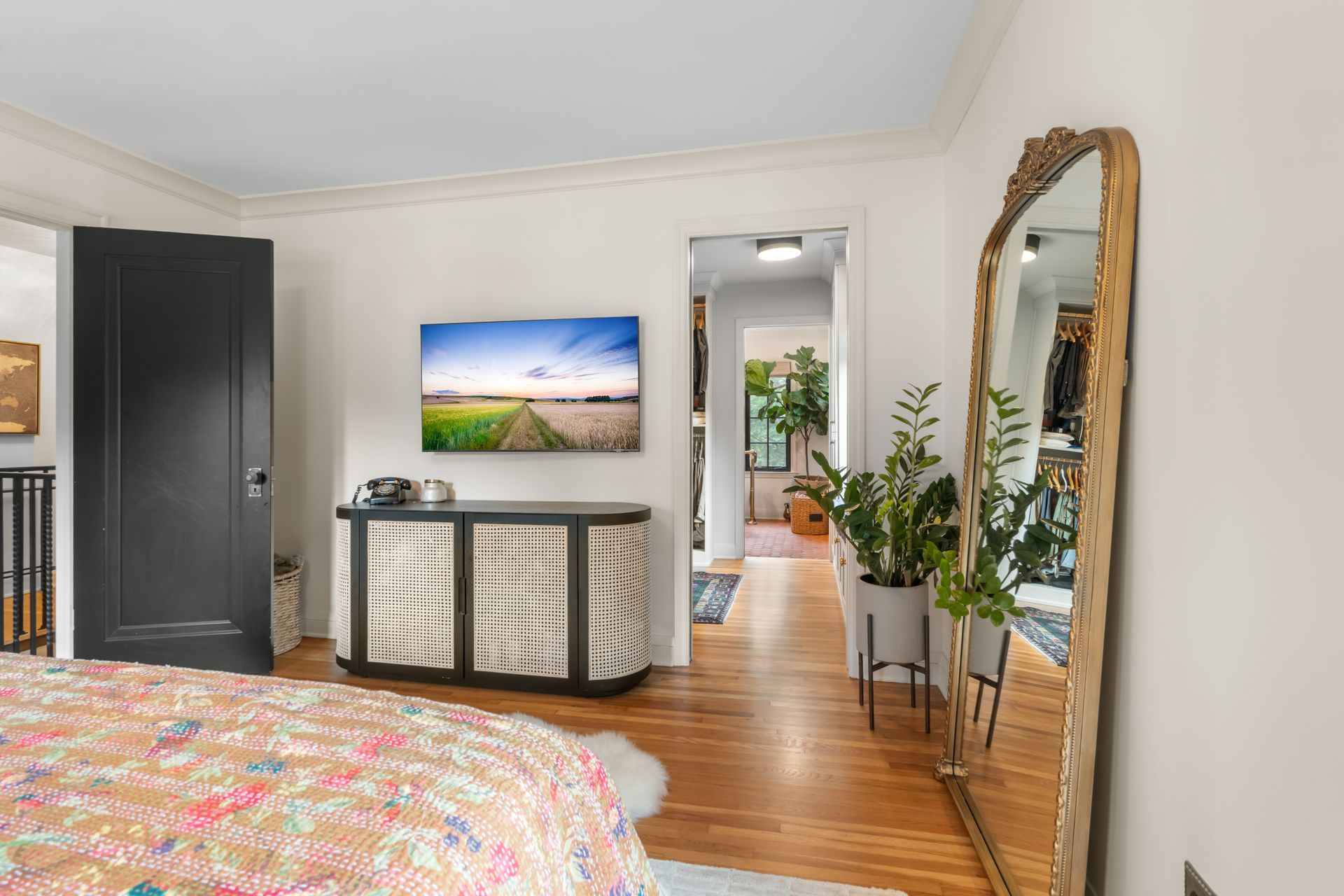 A bedroom featuring a bed with a floral quilt, a decorative black cabinet under a wall-mounted TV, and a gold mirror.
