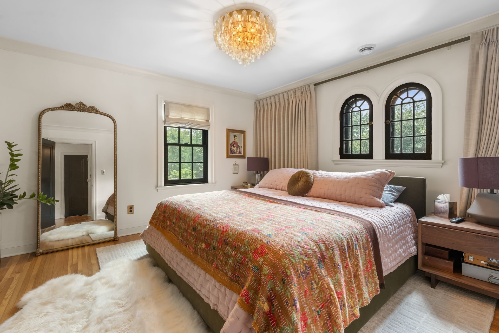 A light-filled bedroom with a king bed, patterned quilt, two arched windows, a full-length mirror, and a gold chandelier.