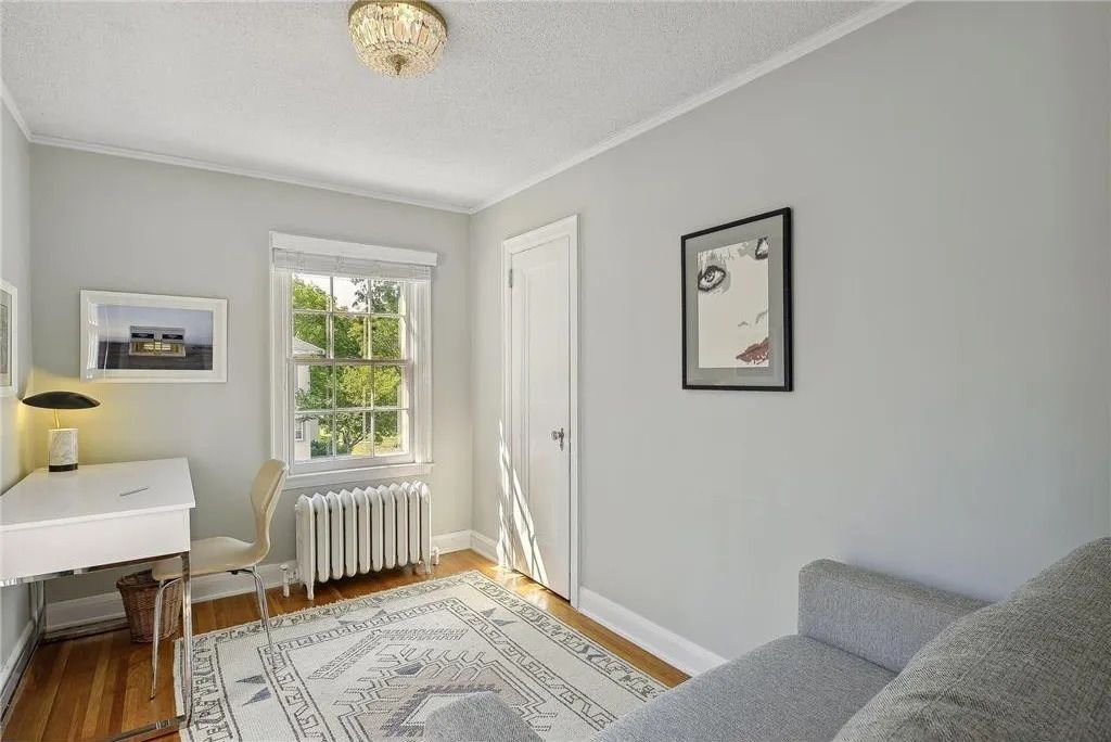 A light-filled home office with a white desk, a patterned rug, a grey sofa, and a framed art print on a light grey wall.
