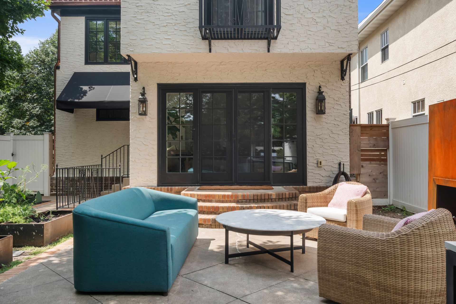 A modern patio with a teal sofa, round coffee table, and wicker chairs in front of a house with black doors and white walls.