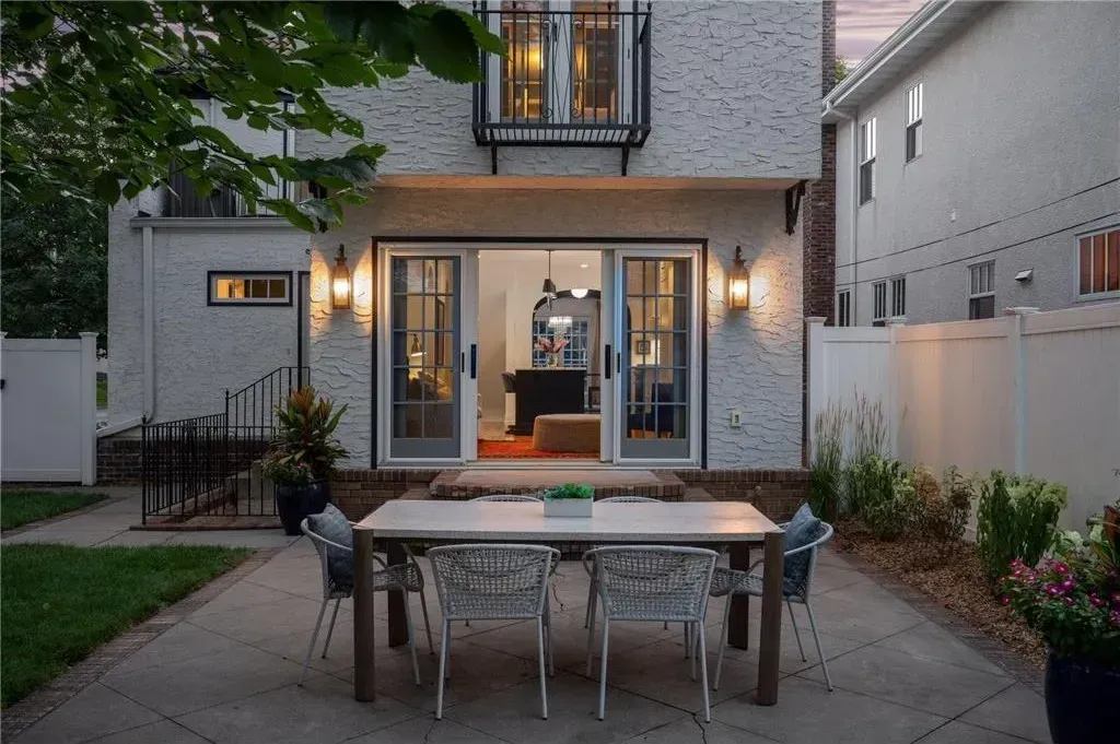 A light-colored patio with a dining table and six chairs sits in front of a house entrance with glass sliding doors.