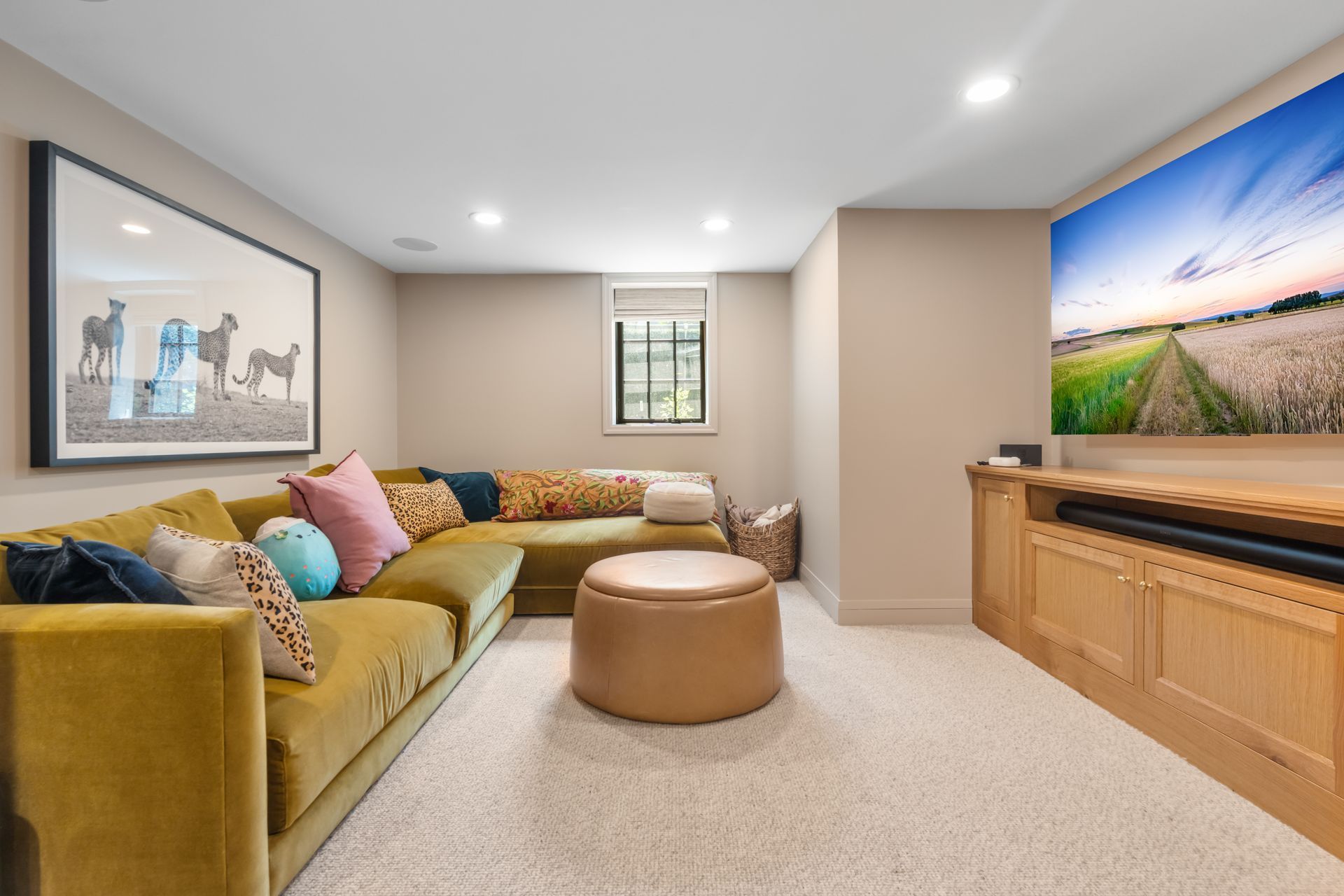 A cozy basement media room with a large mustard sectional sofa, a tan leather ottoman, and a flat-screen TV on wood cabinets.
