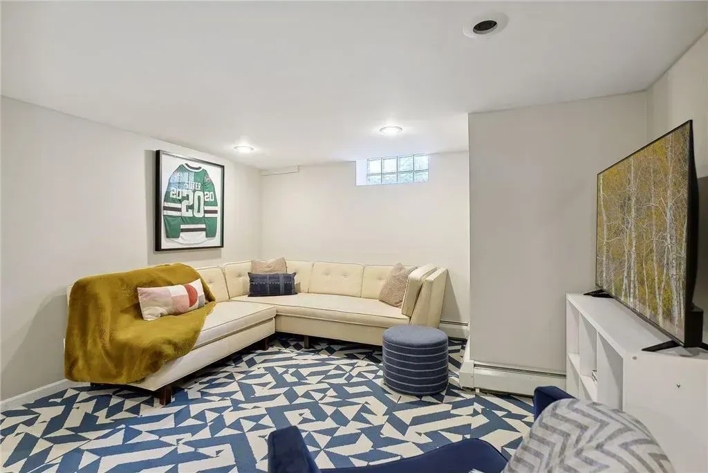 A basement living area with a large cream sectional, blue geometric rug, dark blue ottoman, and a wall-mounted television.