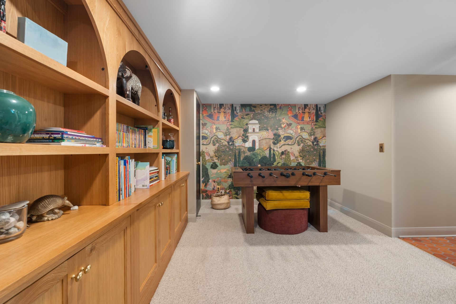 A wood-paneled home library with a foosball table, shelves filled with books, and a scenic wallpapered accent wall.