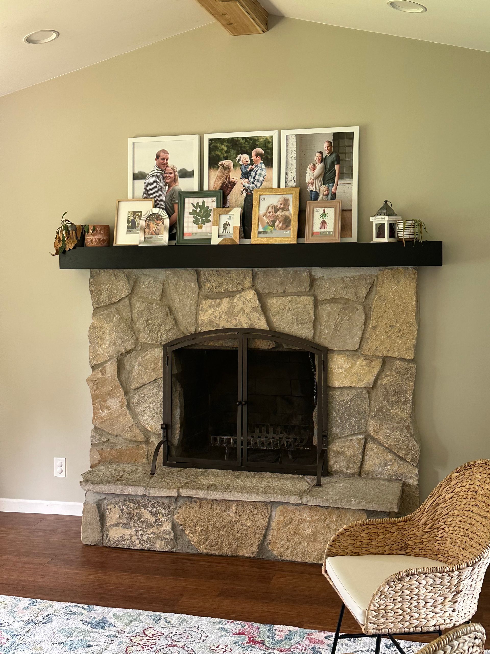 A stone fireplace with a black wooden mantel holding several framed photos and a small white lantern, in a neutral room.