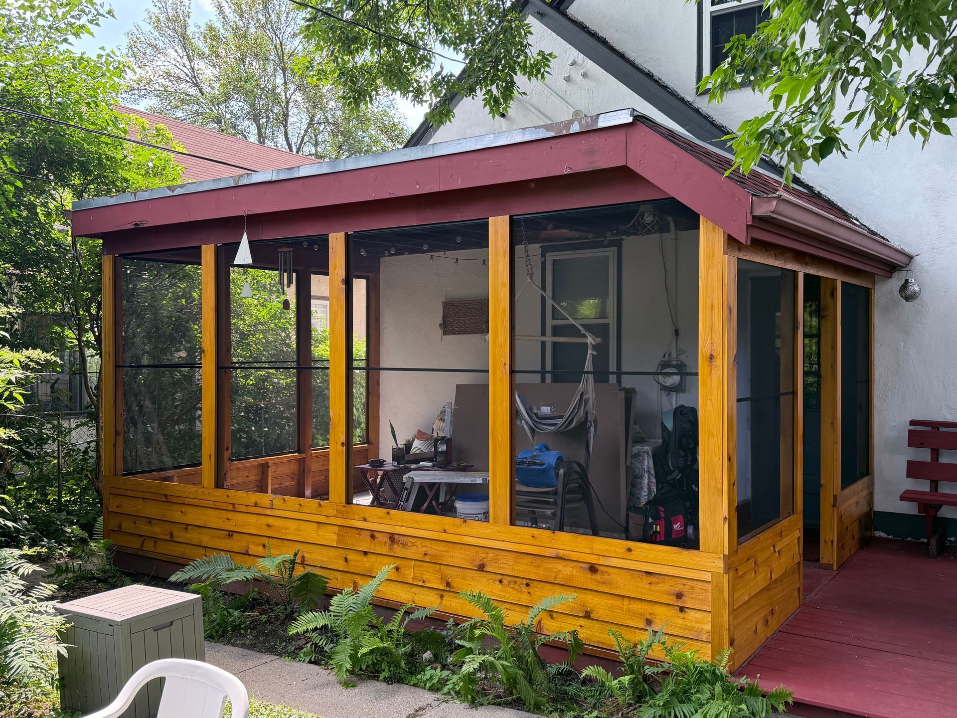 A screened-in porch with natural wood paneling and a dark red trim, attached to the side of a white house.