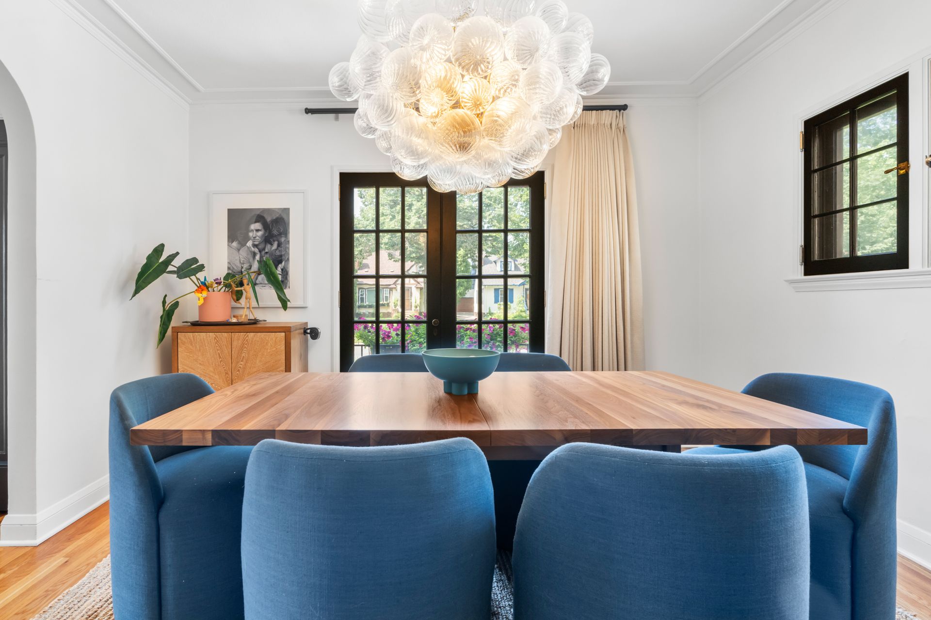 Dining room with a wooden table, blue chairs, a glass bubble chandelier, and a large window looking onto greenery.