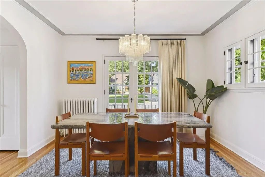 A dining room with a marble-topped table, four wooden chairs, a crystal chandelier, and a houseplant by glass doors.