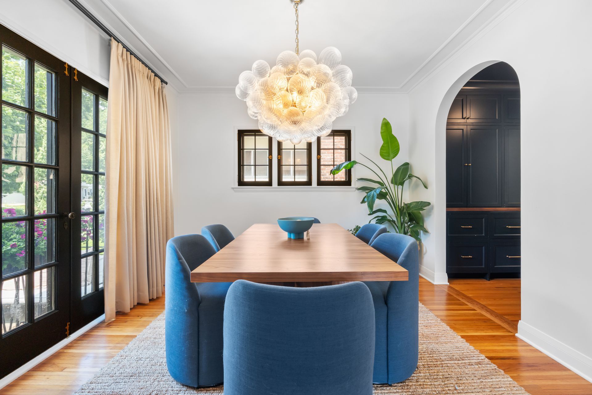 Dining room with a wood table, blue upholstered chairs, modern bubble chandelier, and an arched doorway to a dark study.