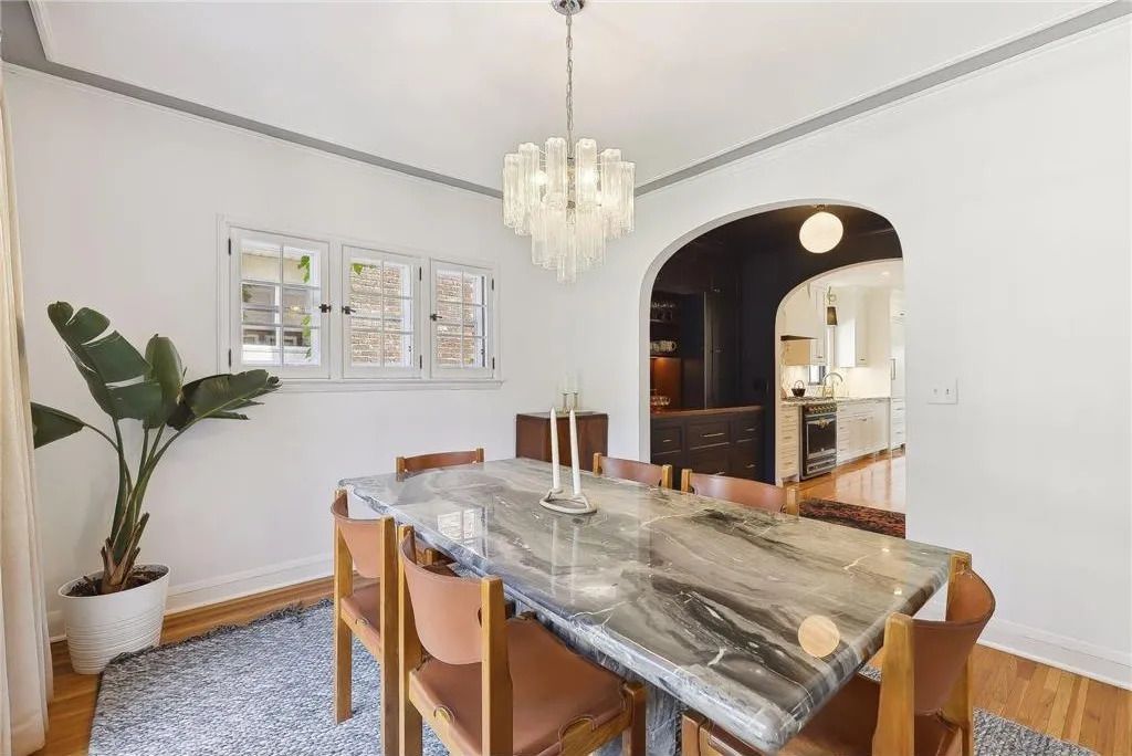 A dining room with a marble table, wooden chairs, a tiered glass chandelier, and a potted plant on a blue patterned rug.