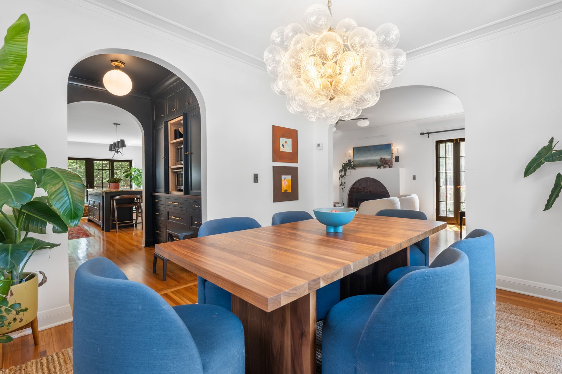 A modern dining room featuring a wooden table with blue chairs, a glass bubble chandelier, and arched doorways.
