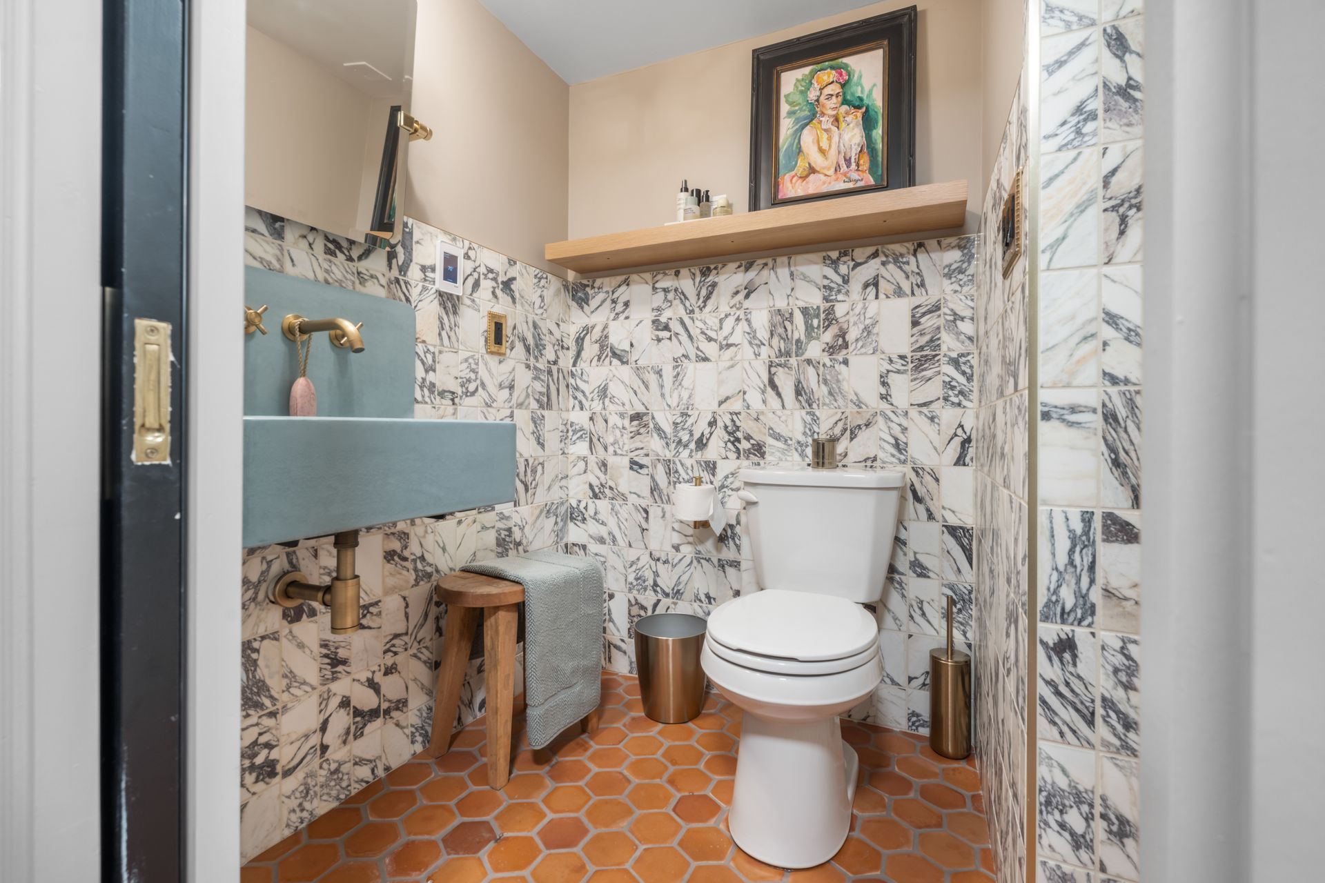 A small bathroom with a blue wall-mounted sink, a white toilet, terracotta hexagonal floor tiles, and black-and-white tile.