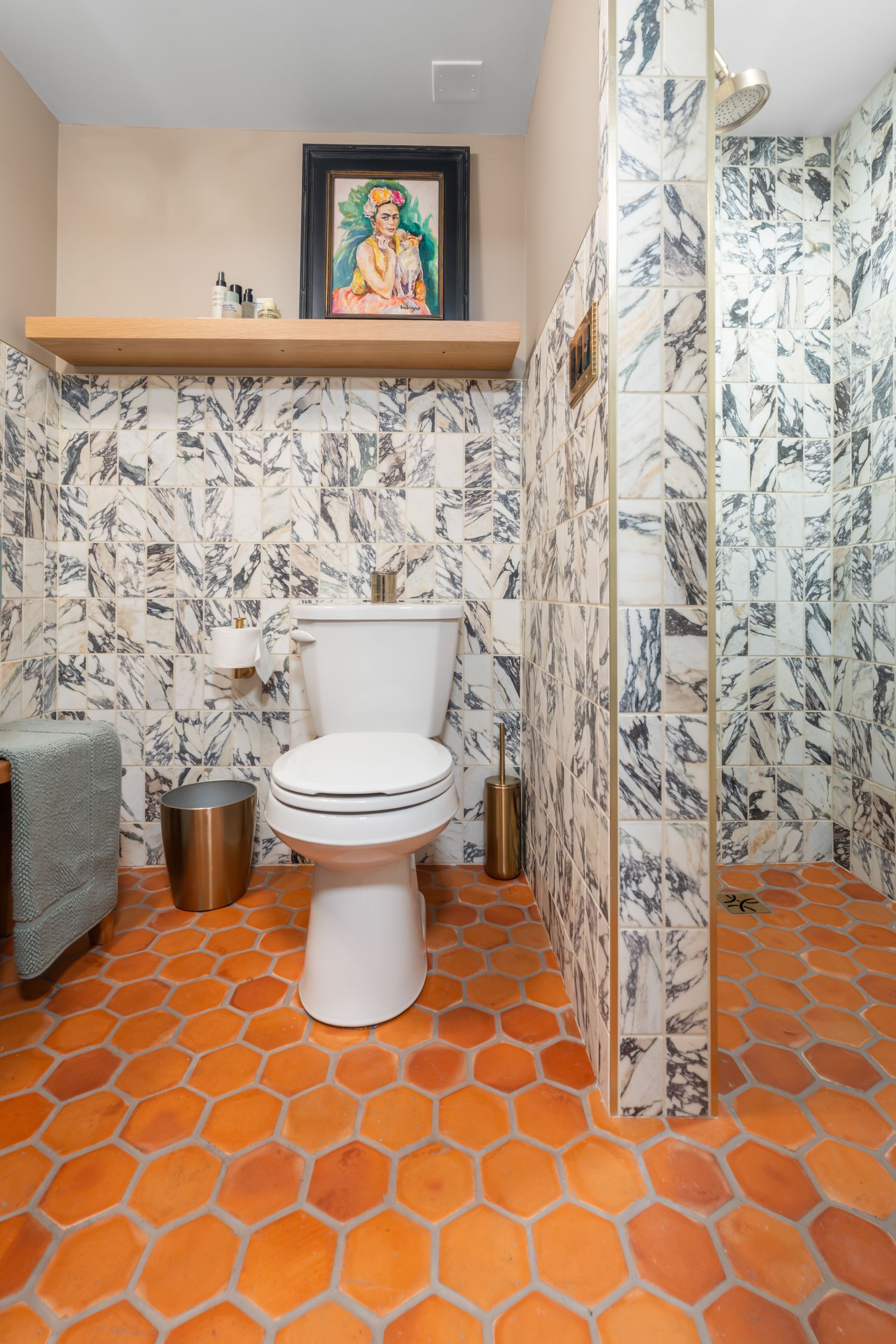 A bathroom with orange hexagonal tile floors, black and white marble-style wall tiles, a toilet, and a wood shelf above.