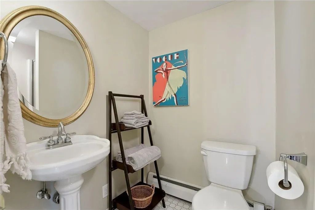 A bright bathroom with a pedestal sink, round gold-framed mirror, wooden towel shelf, toilet, and colorful wall art.