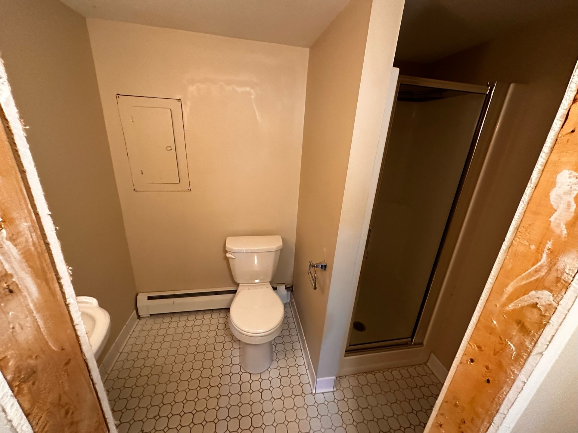 A small, beige-walled bathroom featuring a white toilet, a shower stall, and a section of white tile flooring.