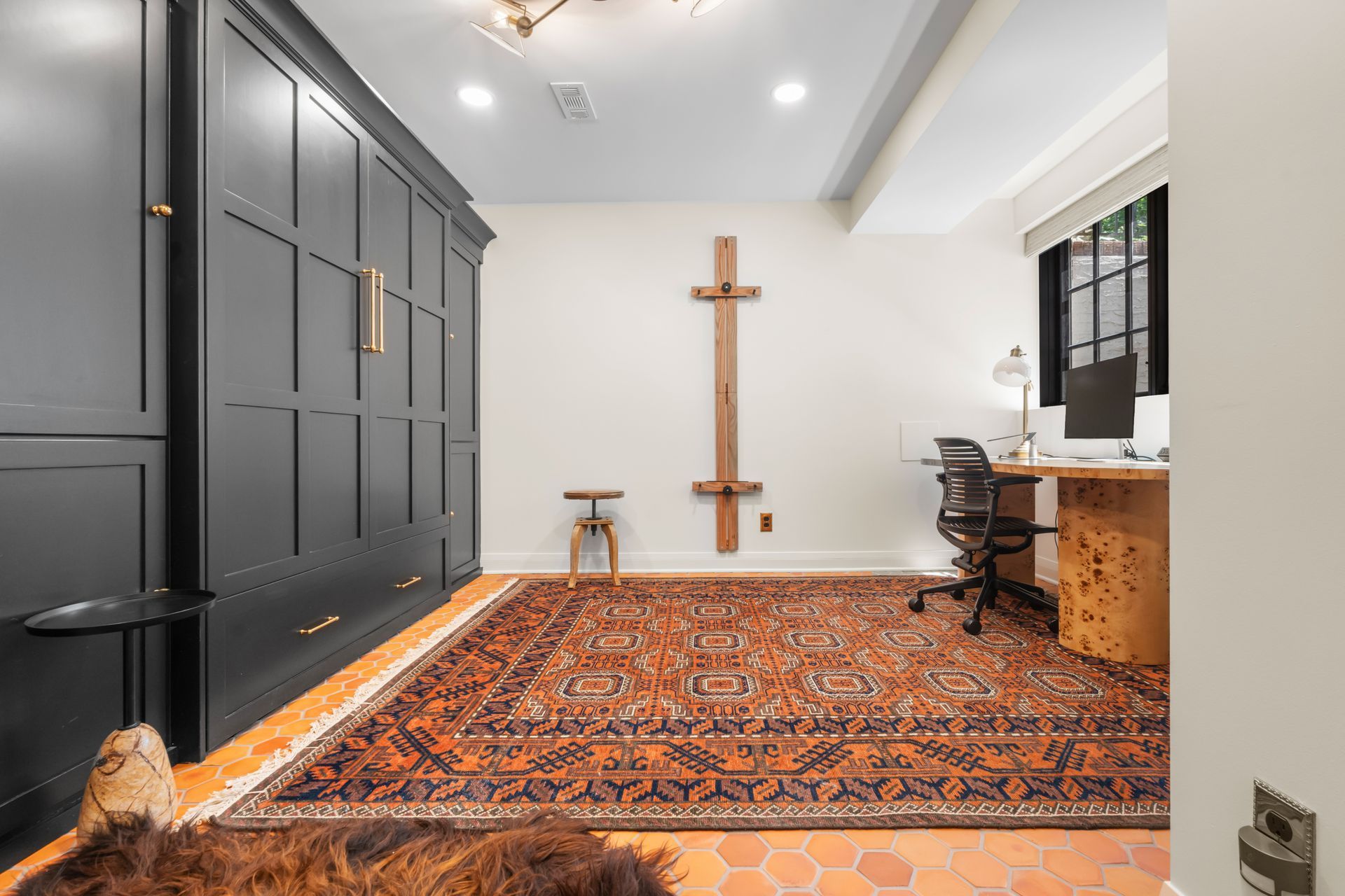 A home office with a dark gray Murphy bed unit, a large patterned area rug, and a desk facing a window with a computer.