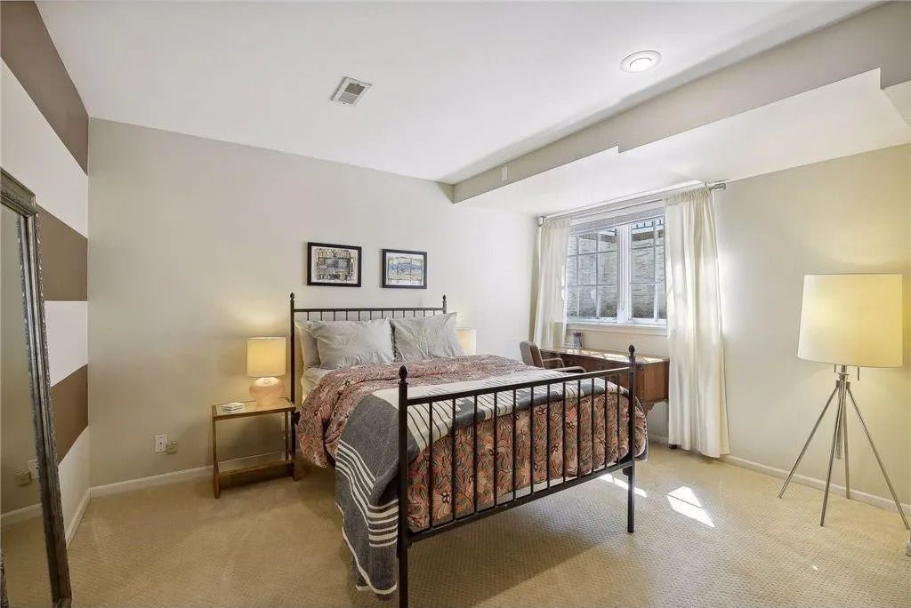 A neutral-toned bedroom with a metal-framed bed, two wall pictures, a side table with a lamp, and a tripod floor lamp.